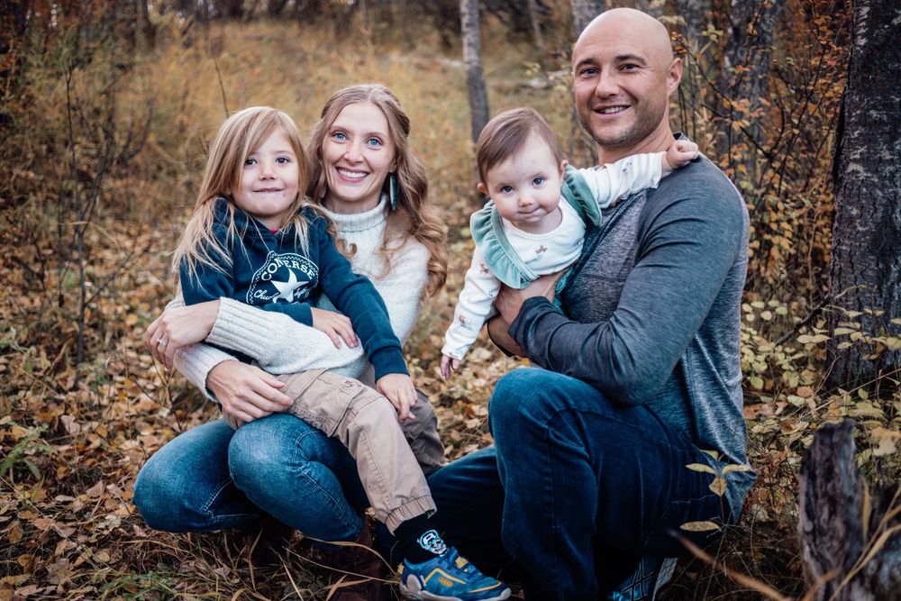 A family portrait captured in an autumn forest setting with fallen leaves and muted natural colors.