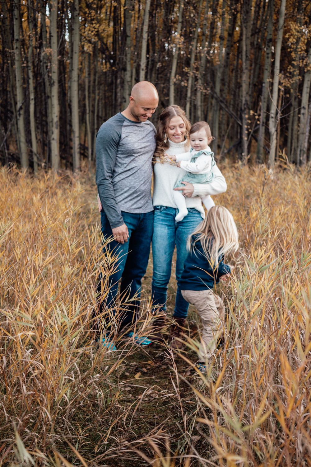 A family stands together in tall autumn grass near a forest of bare trees on a fall day.