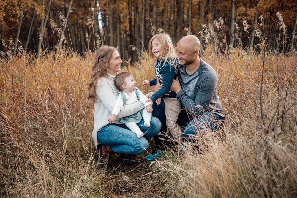 A family sits together in tall autumn grass with bare trees and warm fall colors in the background.