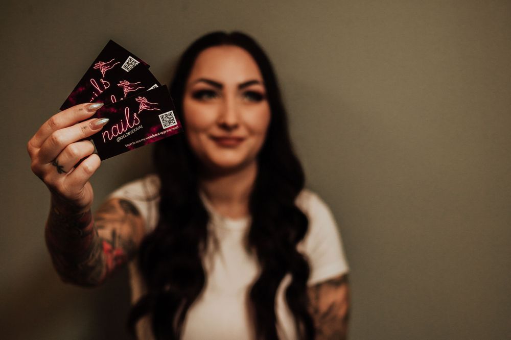 Someone with long dark wavy hair and tattoos holds up what appears to be a gift card while wearing a white shirt.