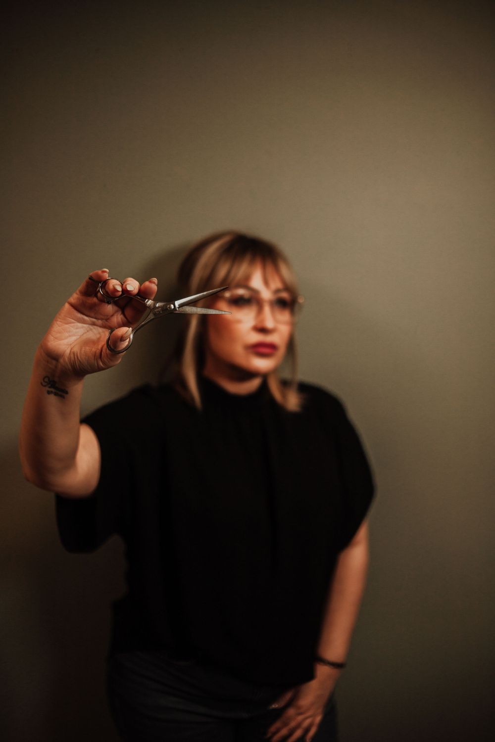 Professional wearing black clothing holding up scissors against moody brown background in artistic portrait.