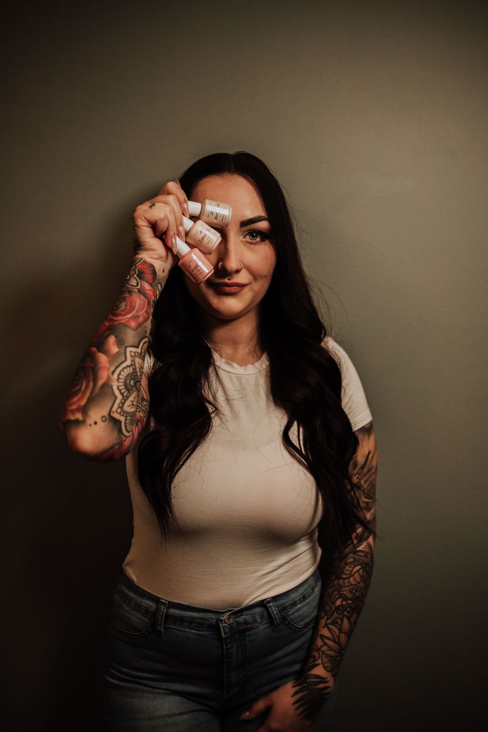 Woman with long dark hair wearing white t-shirt and jeans shows off intricate sleeve tattoo designs against dark background.