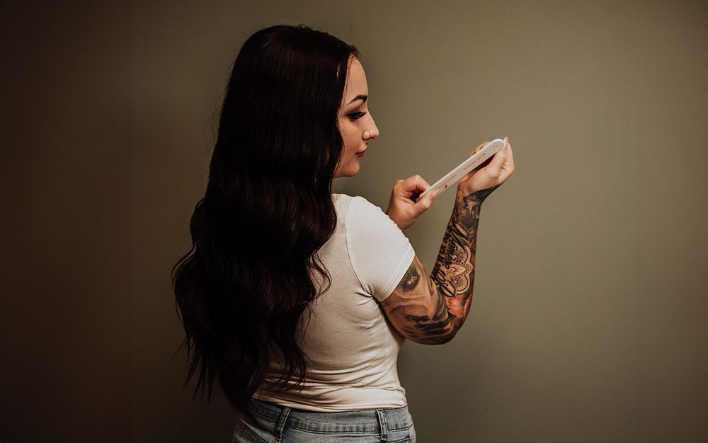 Side view of a woman with dark hair wearing a white crop top and jeans showing her tattooed arm against a brown background.