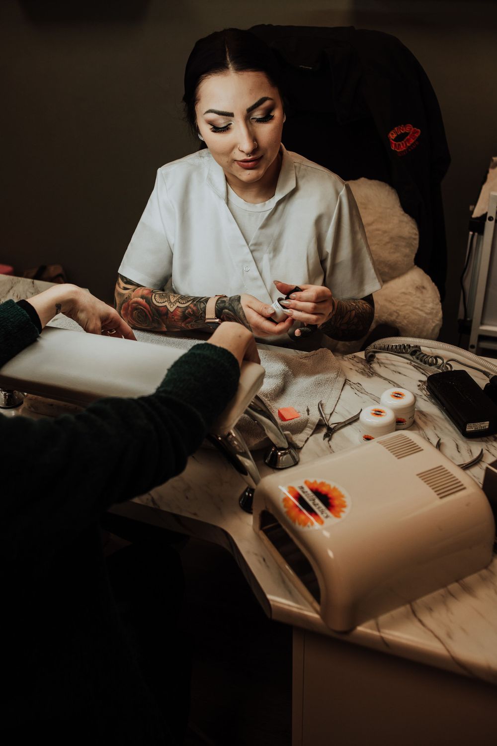 Professional nail technician with sleeve tattoos working on client's manicure at salon workstation with tools and equipment.