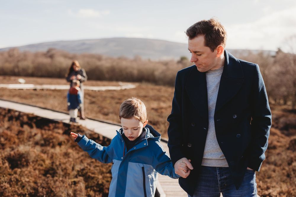 A father holds his young son's hand on a wooden boardwalk trail through autumn moorland, with hills in the background.