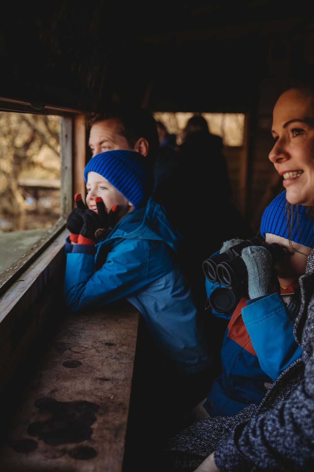 A woman and child in blue winter jackets and hats look out a wooden cabin window on a cold day.