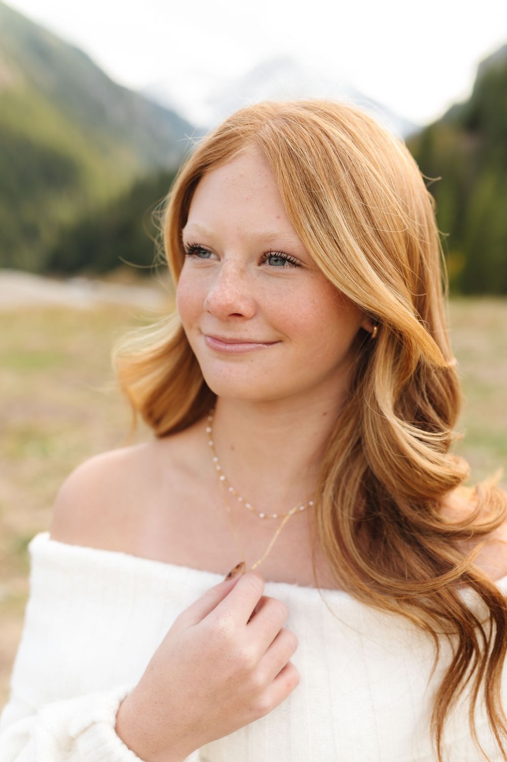 Young woman with long red hair and freckles wearing an off-shoulder white top and delicate necklace in a mountain meadow.