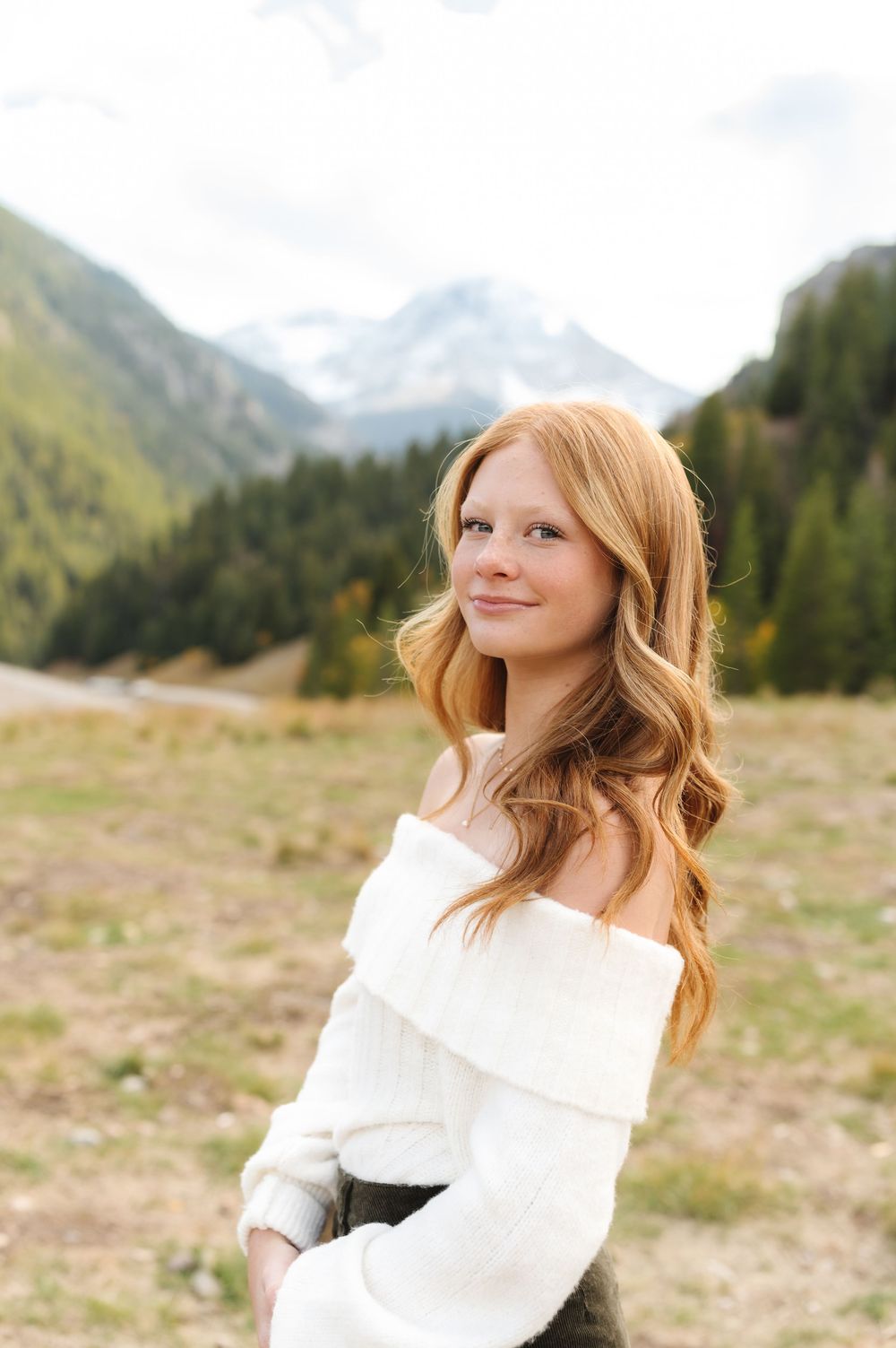 Young woman with long red hair in white off-shoulder sweater smiling outdoors with mountain scenery behind her.