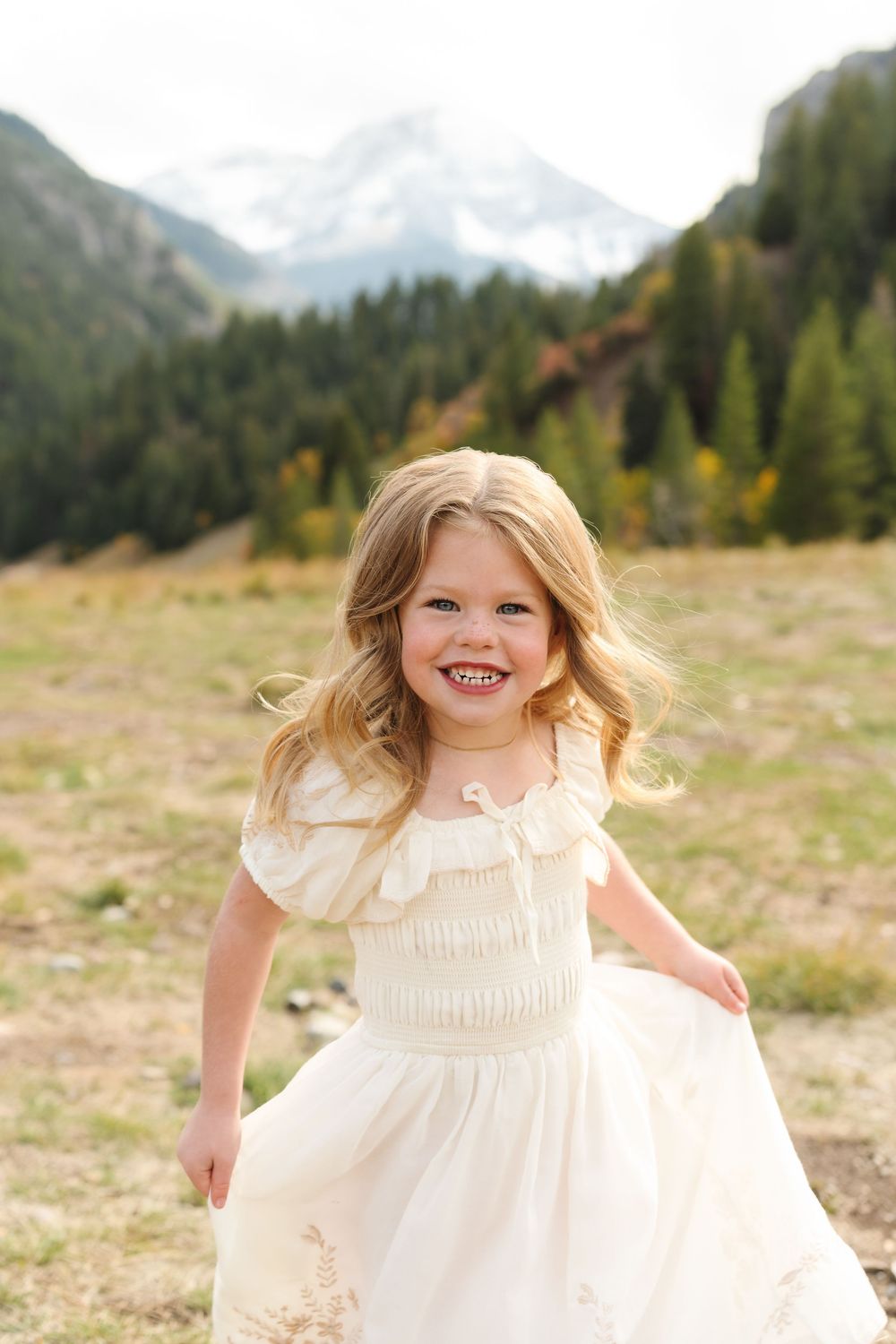 A smiling little girl in a white dress twirls in a mountain meadow with snow-capped peaks and pine trees behind her.