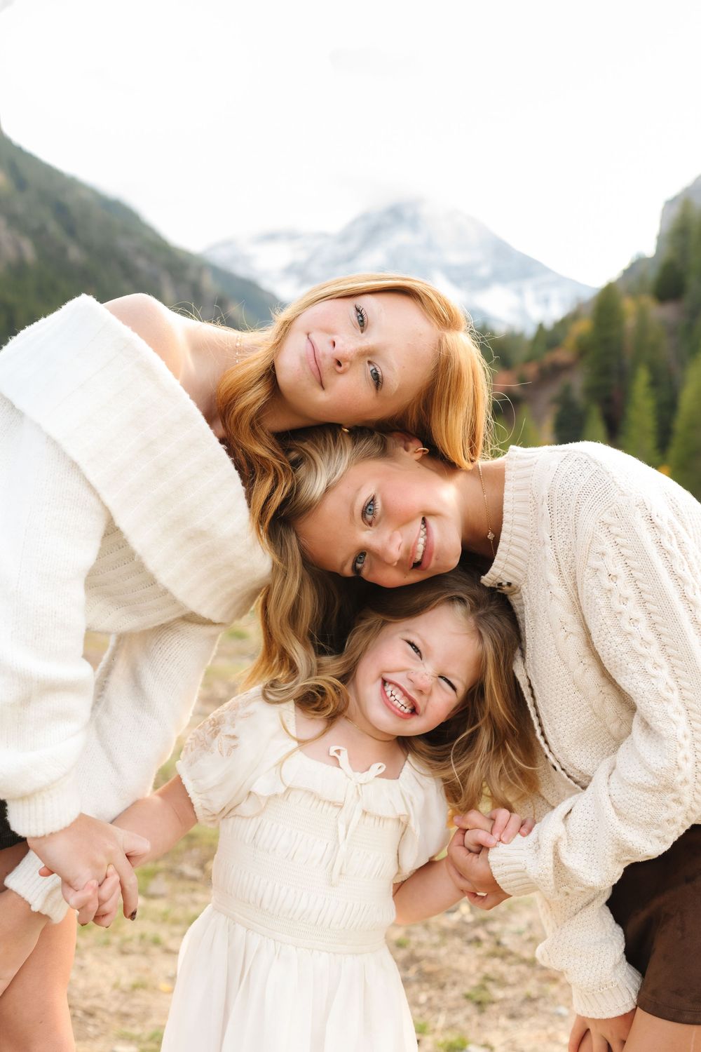 Three smiling girls in white outfits pose together outdoors with snow-capped mountains in the background.
