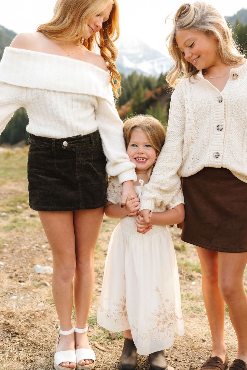 Two blonde women and a smiling little girl in cream and brown outfits hold hands outdoors near mountains.