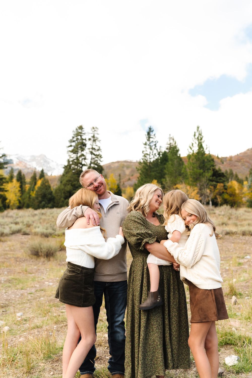 Family hugging outdoors in mountain meadow with fall foliage and evergreen trees in background.