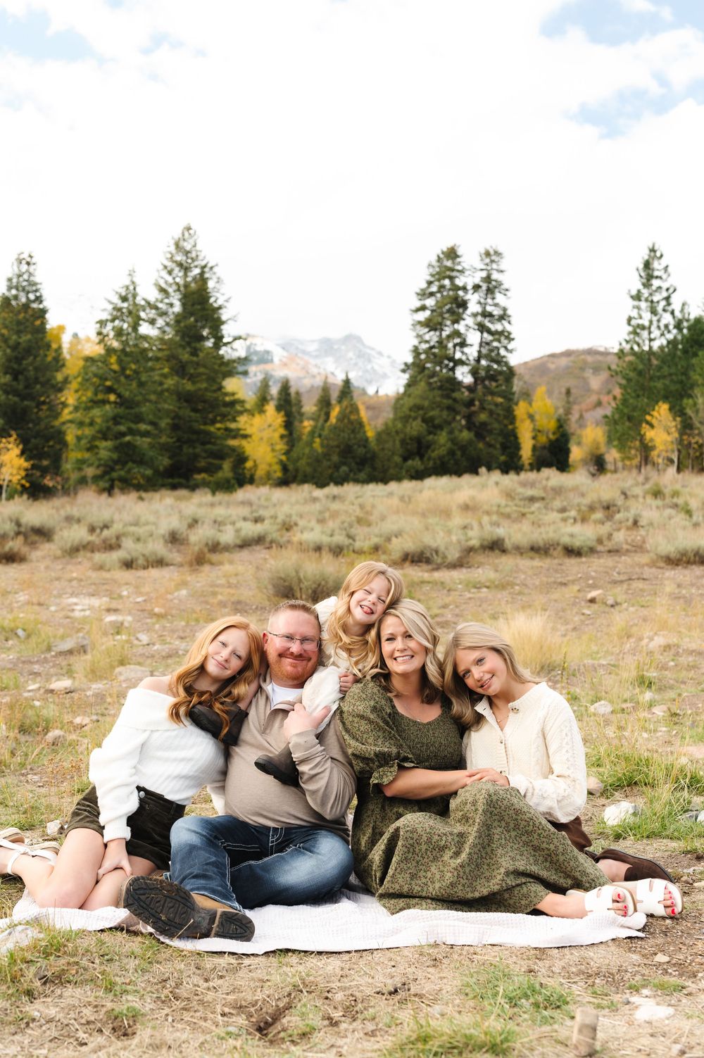 Family of five smiling together on a blanket in an autumn mountain meadow with evergreen trees.