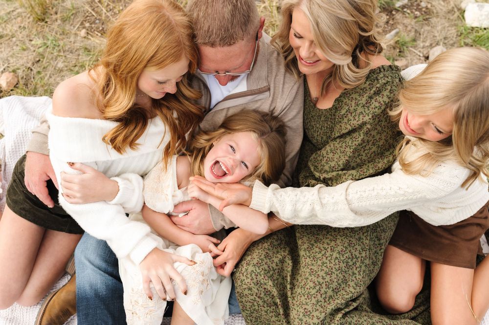 A joyful family of five laughs together outdoors, with parents and three daughters sharing a warm, playful embrace.