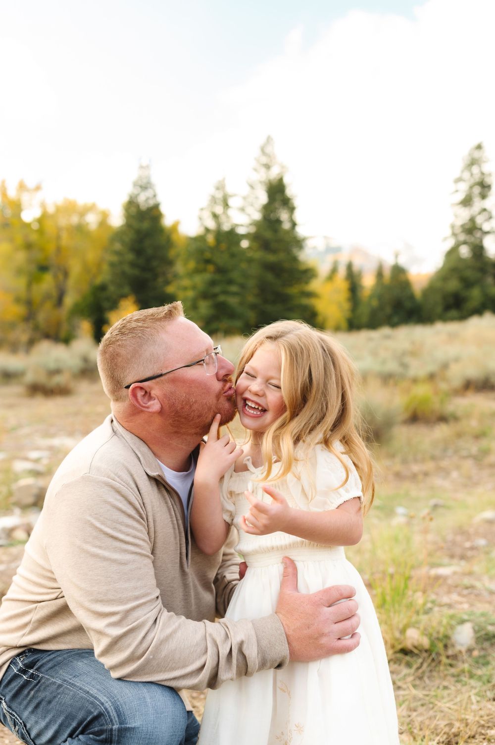 A father kisses his laughing blonde daughter outdoors in a golden autumn meadow with evergreen trees behind them.