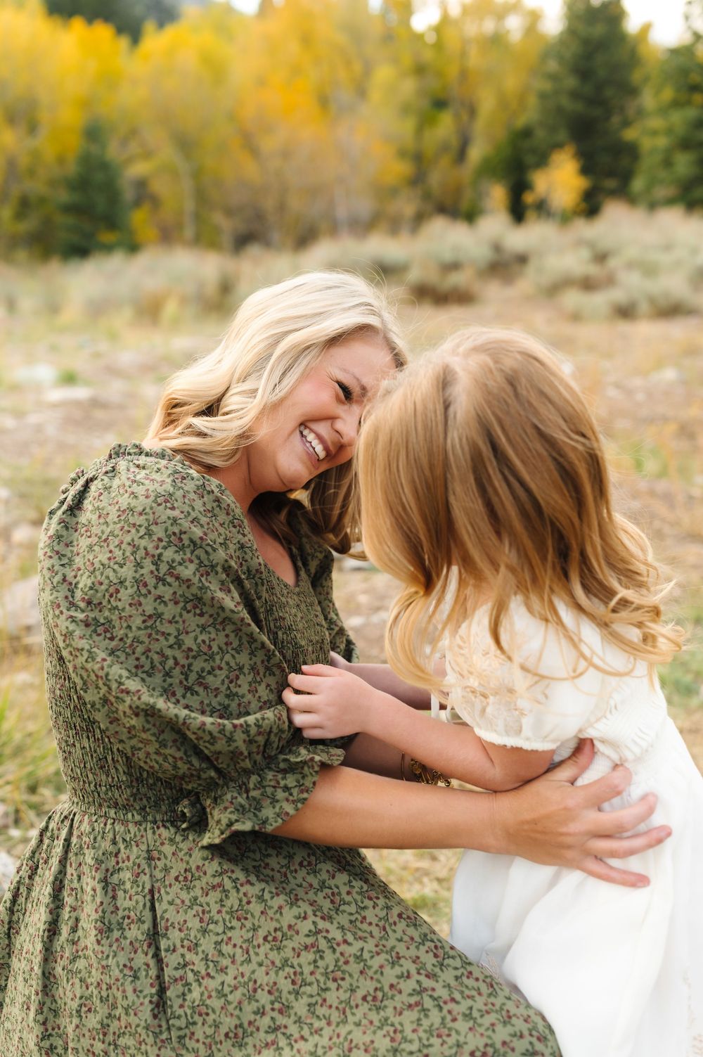 A laughing blonde woman in a green floral dress hugs a young girl in white outdoors among autumn trees.