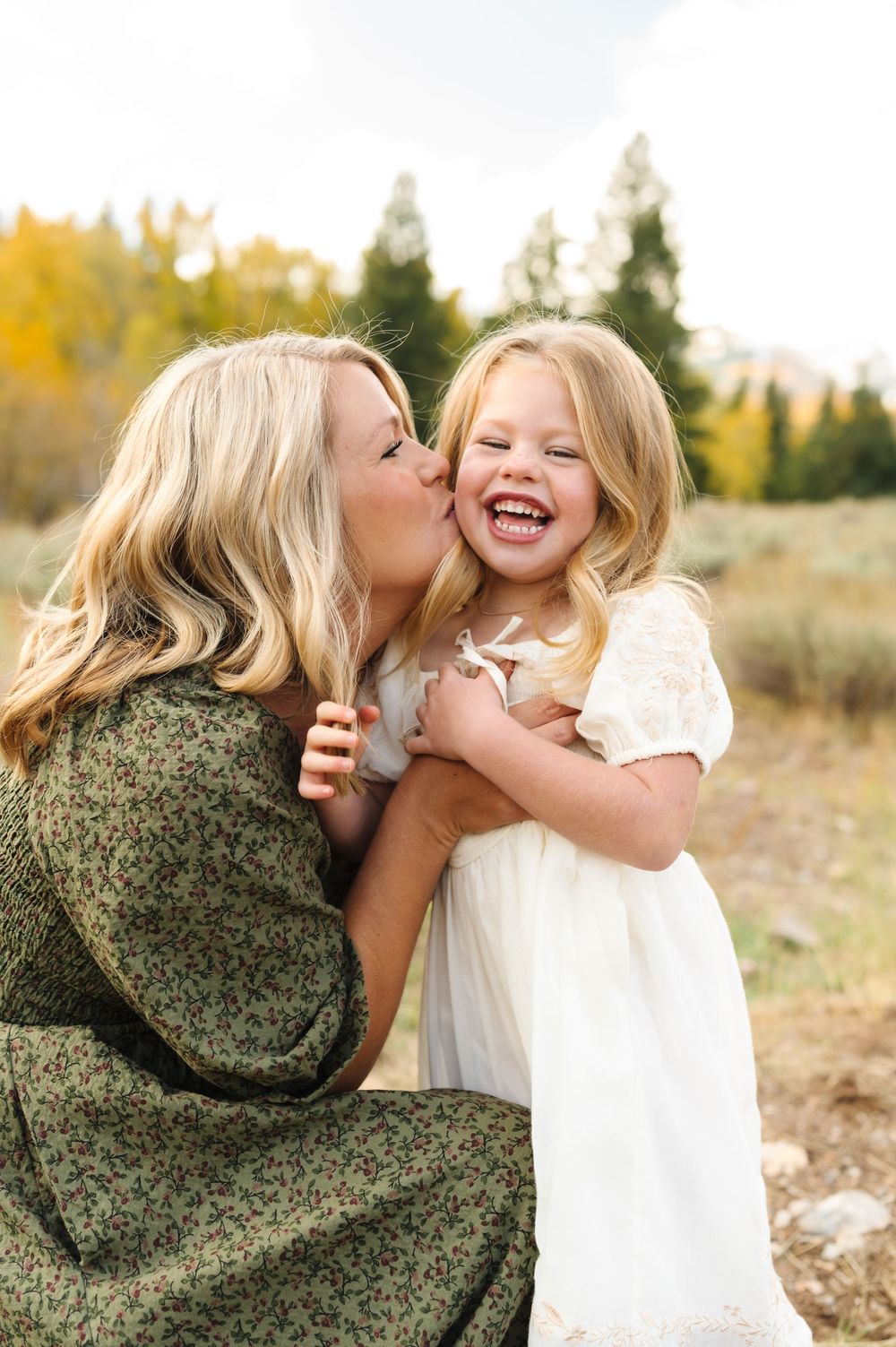 A blonde mother in a green floral dress kisses her laughing young daughter in a white dress outdoors in autumn.