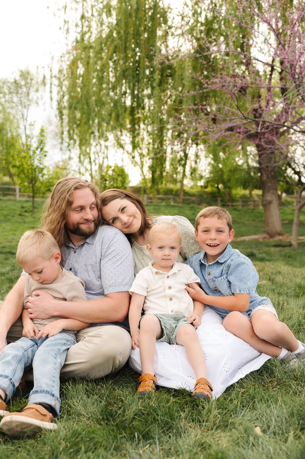 A happy family of five sits together on grass in a lush green park with willow and blooming trees.