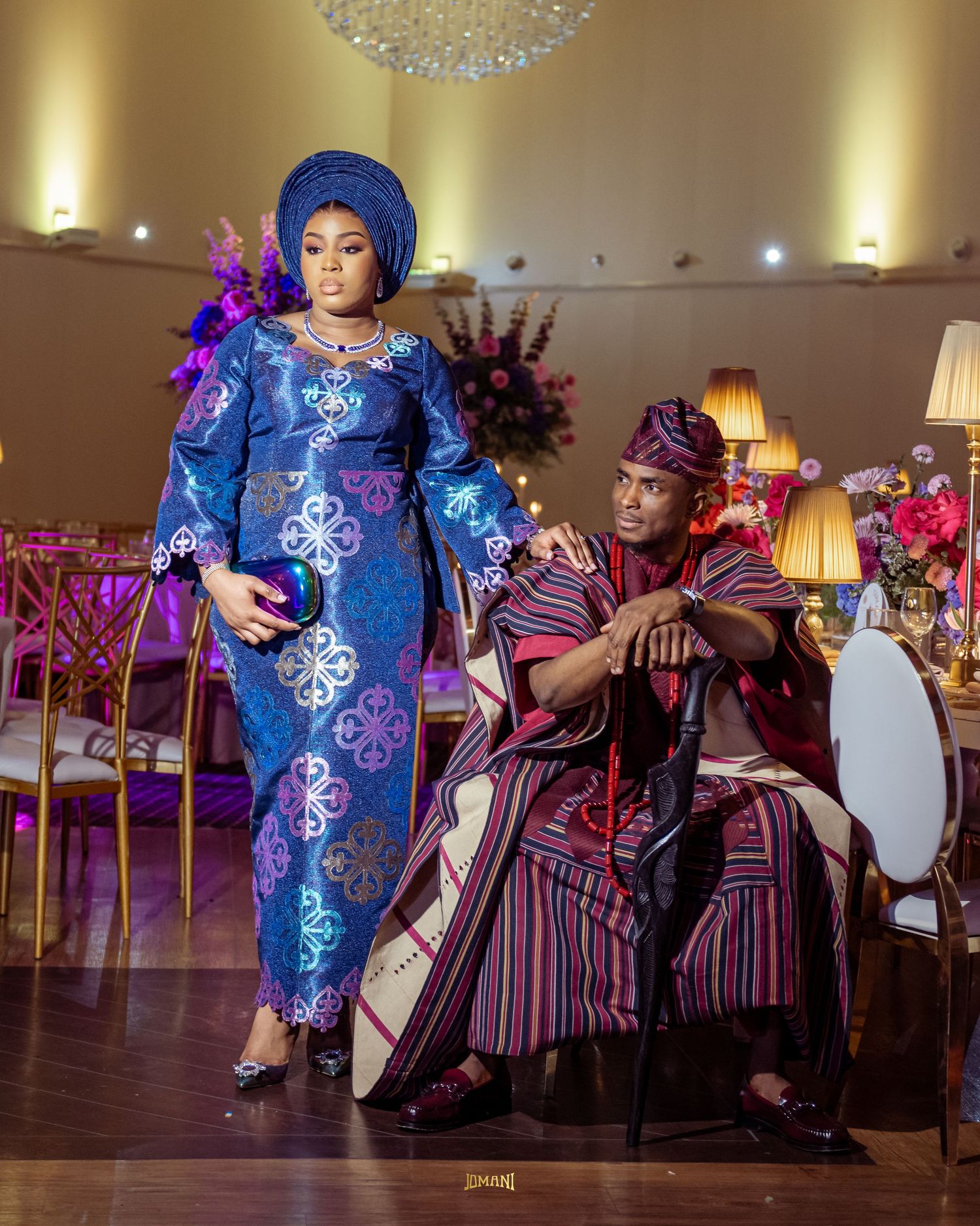 Couple in elegant Nigerian traditional attire pose in decorated wedding reception hall with floral arrangements.
