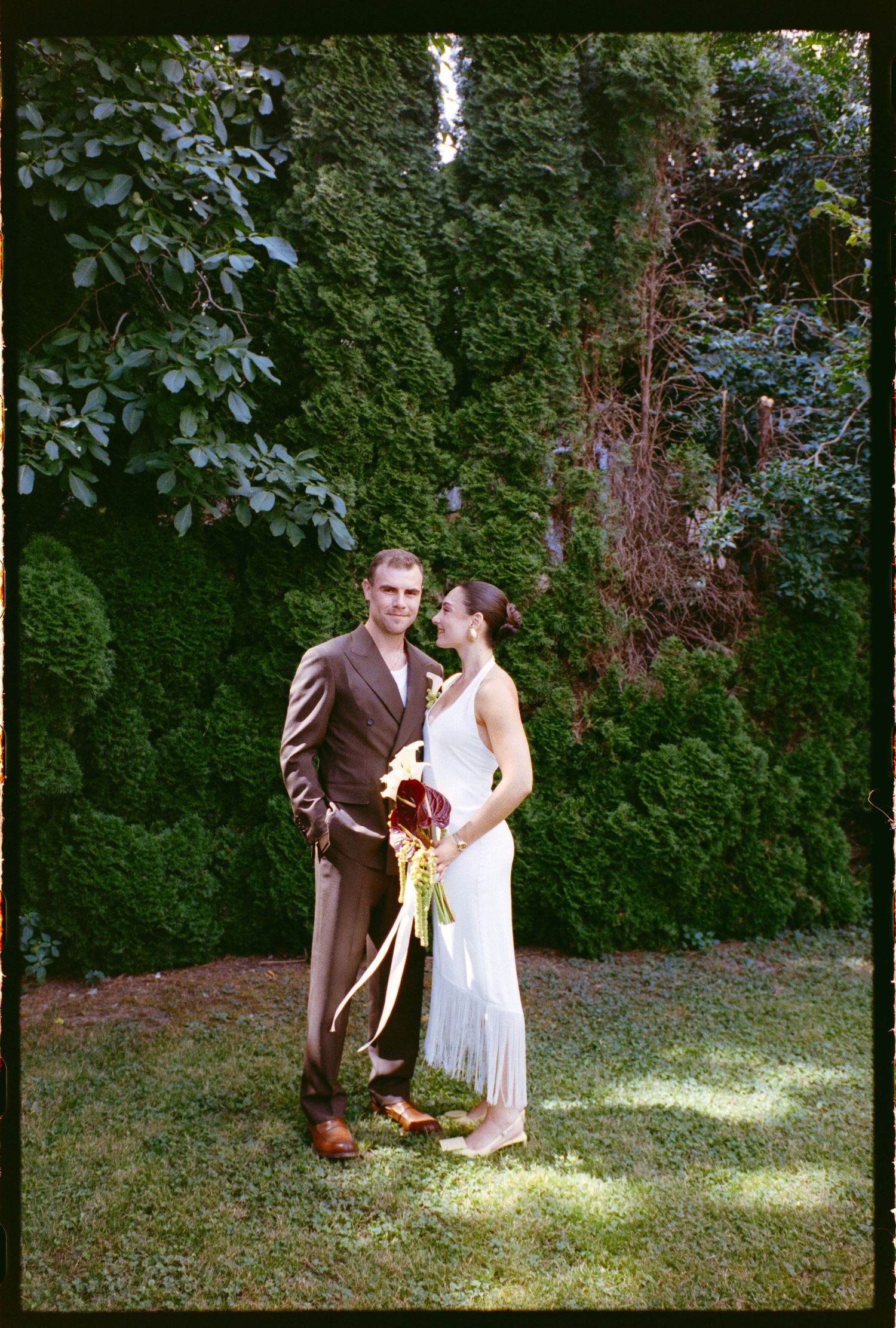 Bride and groom pose together in lush garden setting, she holds red bouquet in white dress, he wears brown suit.