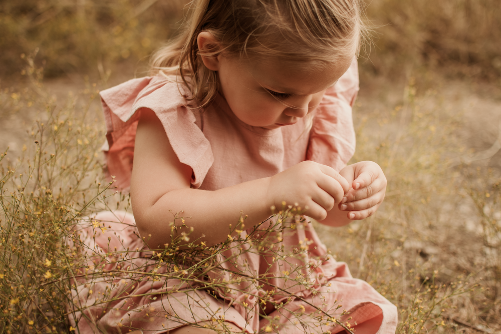 A young girl in a pink ruffle dress crouches in a field, carefully examining small wildflowers.