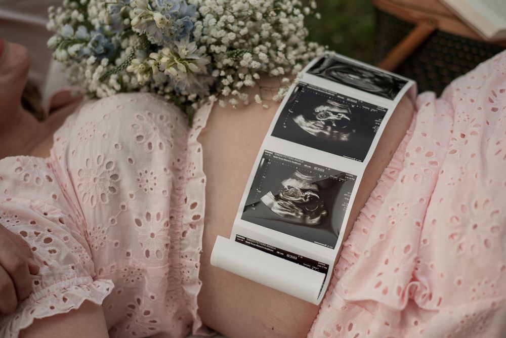 Pregnant woman in pink dress holding ultrasound sonogram photos with baby's breath flowers during maternity photoshoot.