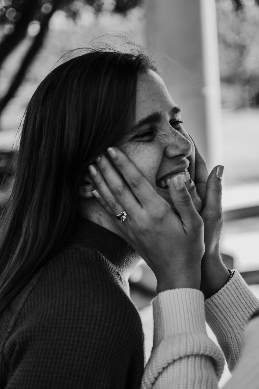 Black and white photo of a laughing woman holding her face with both hands, wearing a ring, outdoors.