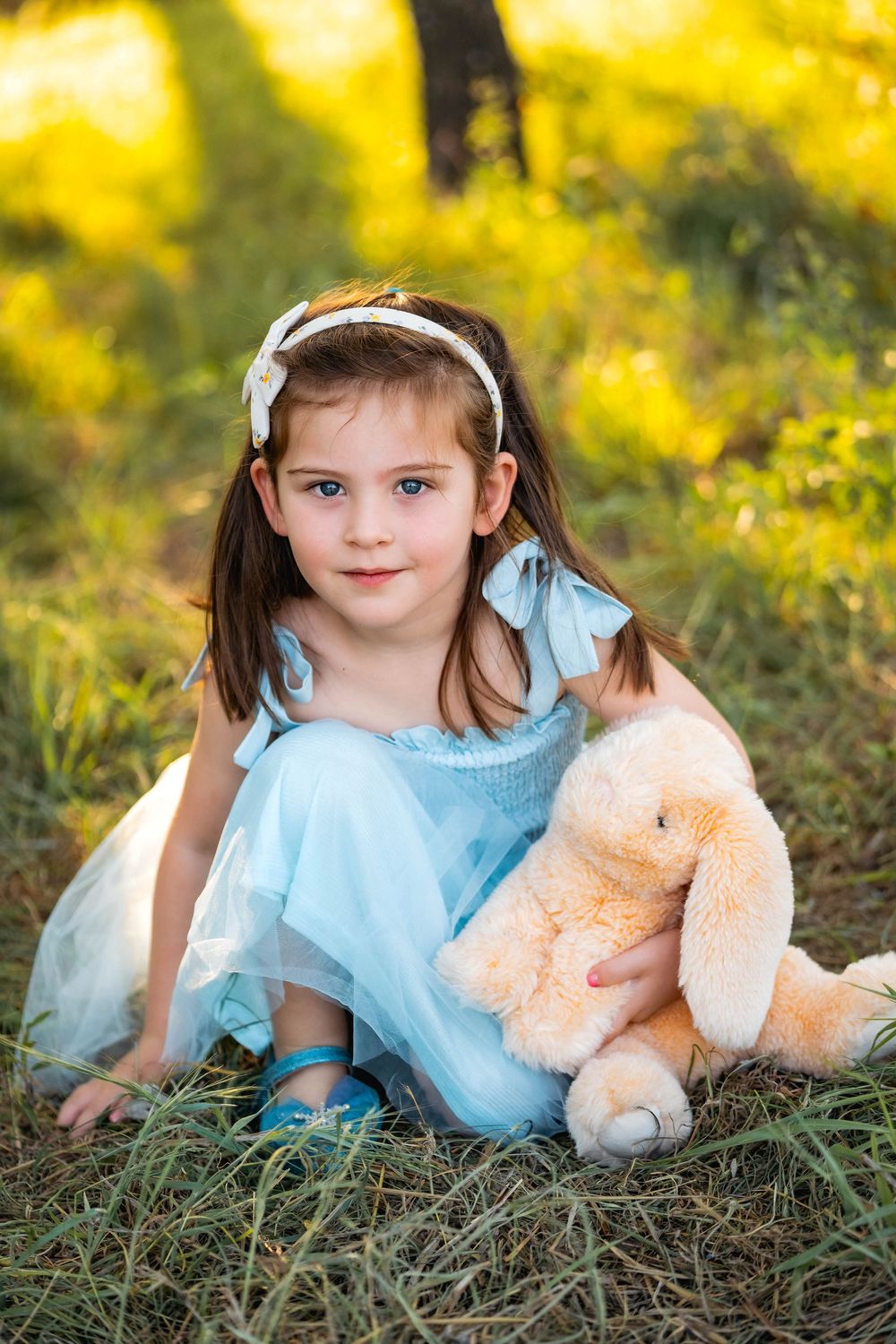 A child in a blue dress lies in grass with a plush teddy bear surrounded by yellow flowers in golden sunlight.