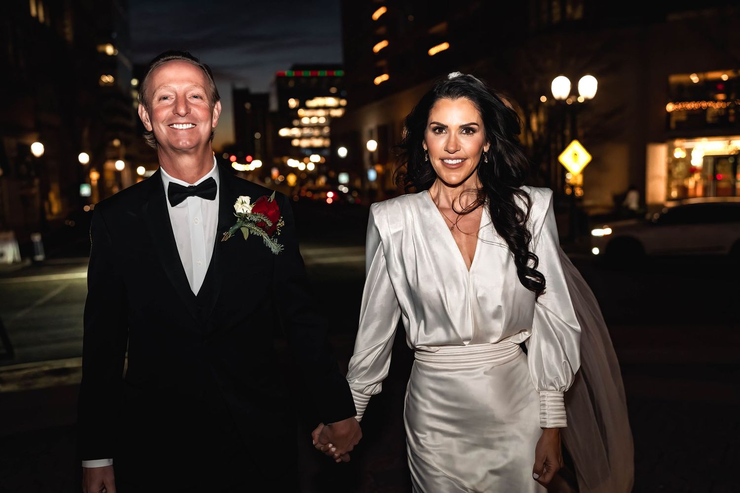 Couple in formal evening attire walking down city street at night with illuminated buildings in background.