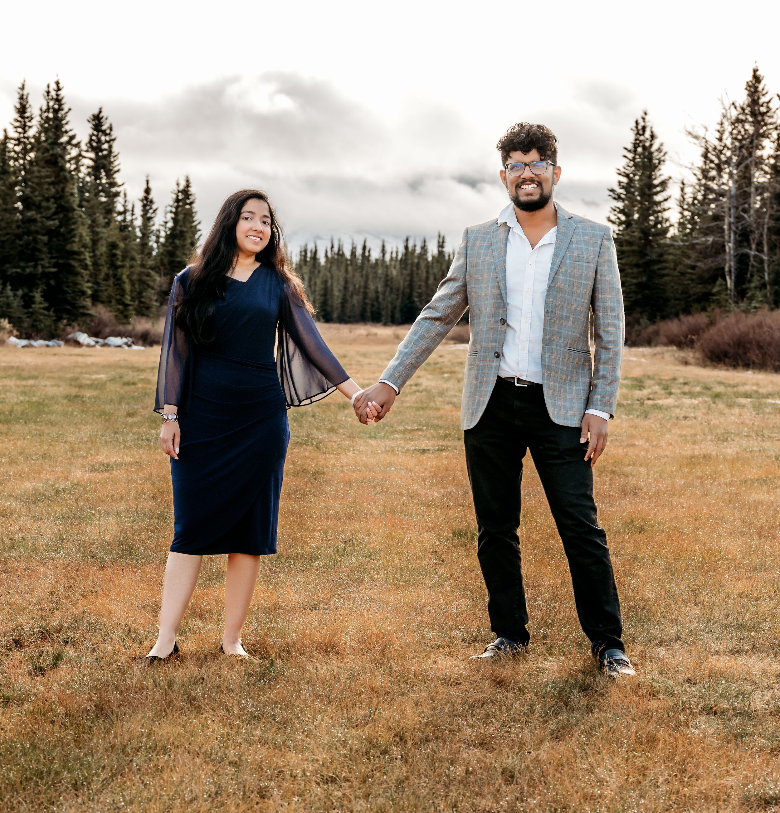 A couple holds hands while posing in a mountain meadow with evergreen trees in the background.