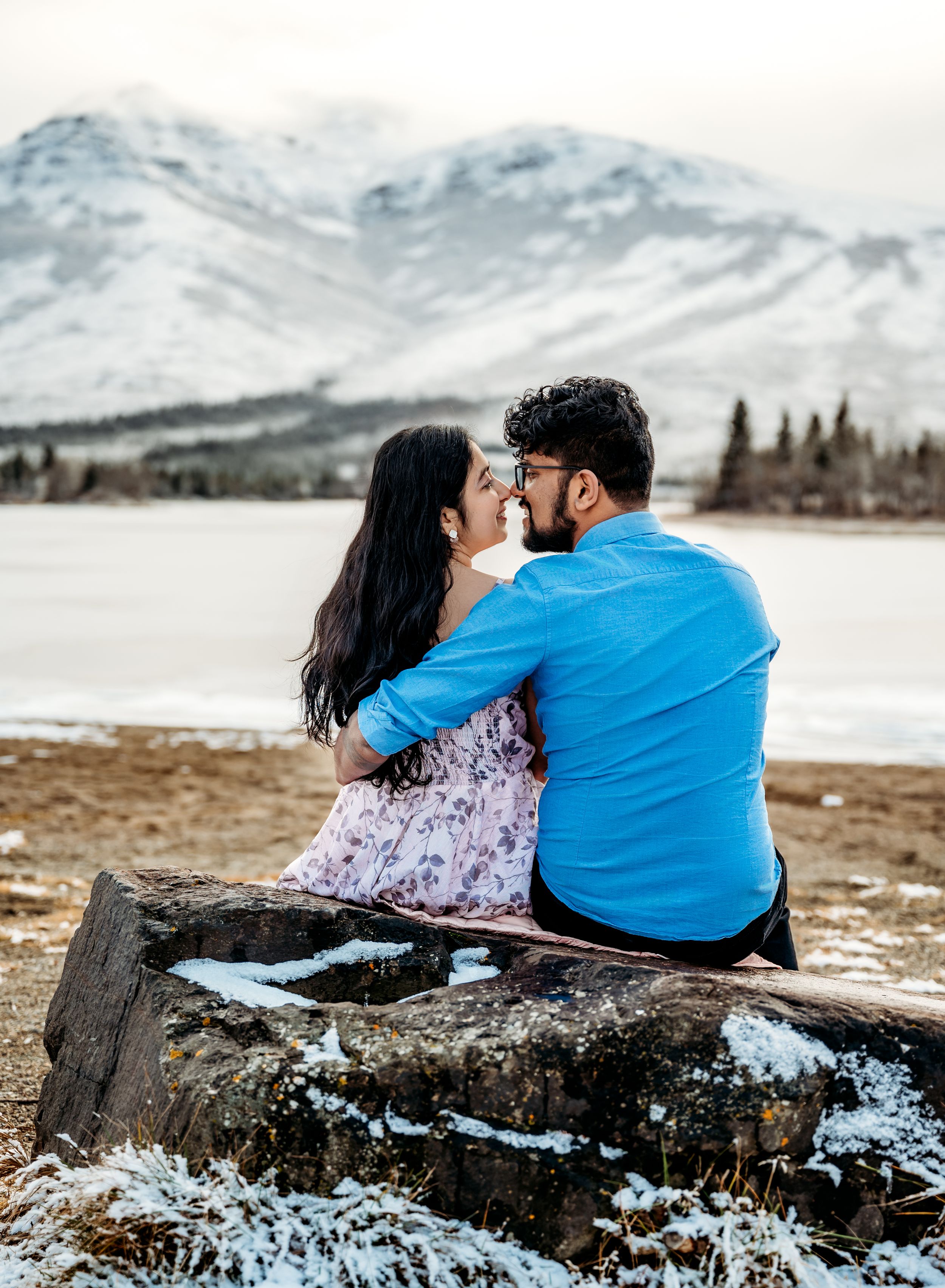 A couple sits together on a rock overlooking a snow-capped mountain landscape.