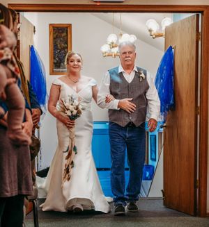 A bride in white gown walks down church aisle accompanied during ceremony.