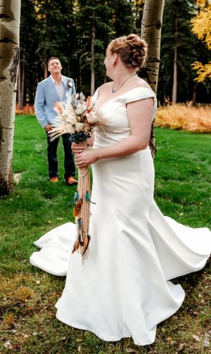 A wedding scene outdoors with a bride in a flowing white dress holding a dried floral bouquet near birch trees.