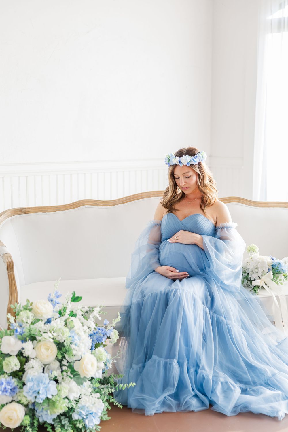 Ethereal maternity photoshoot featuring a woman in flowing blue tulle gown posing elegantly with floral arrangements.