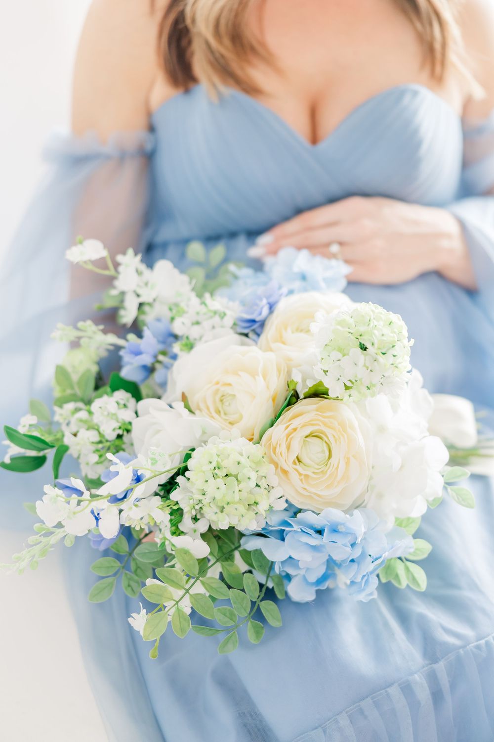 Delicate arrangement of white roses and blue hydrangeas with greenery against soft blue fabric.