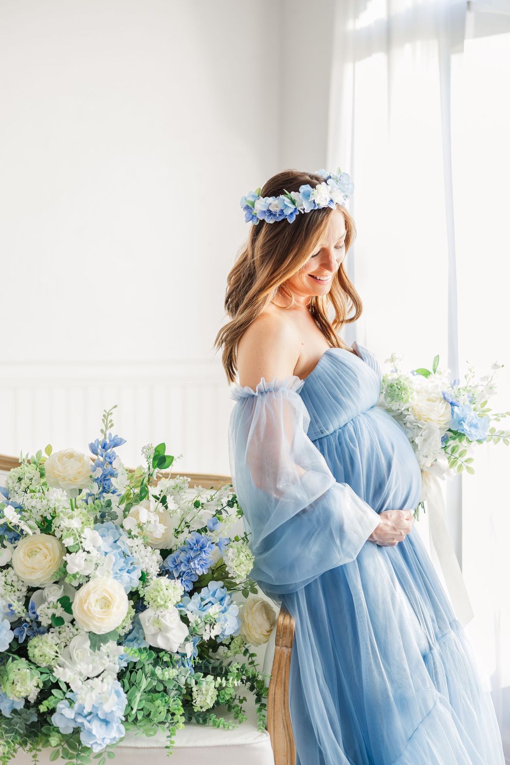 Romantic maternity session with blue gown and white floral backdrop in natural window light.
