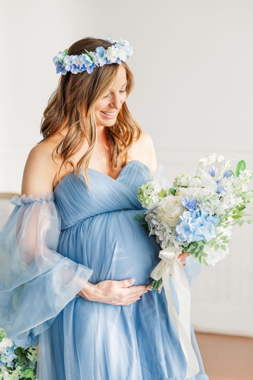 Elegant maternity portraits with blue floral crown and cascading bouquet in white studio setting.