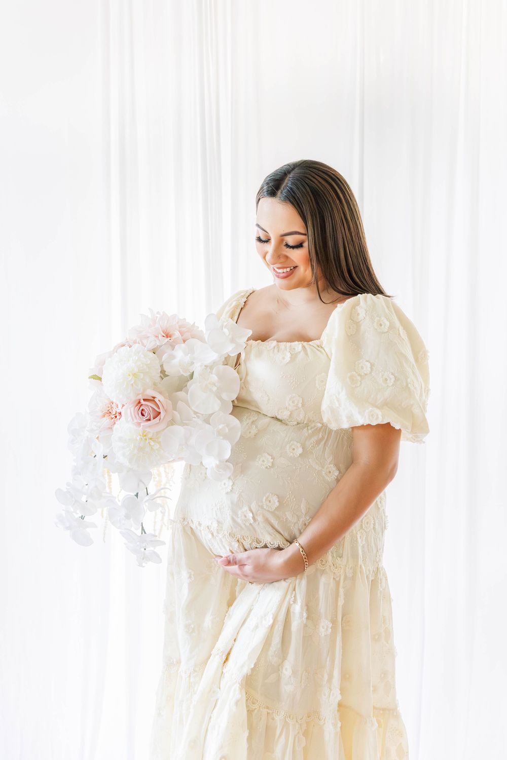 A pregnant woman in a white lace dress poses with flowers during a studio maternity session.