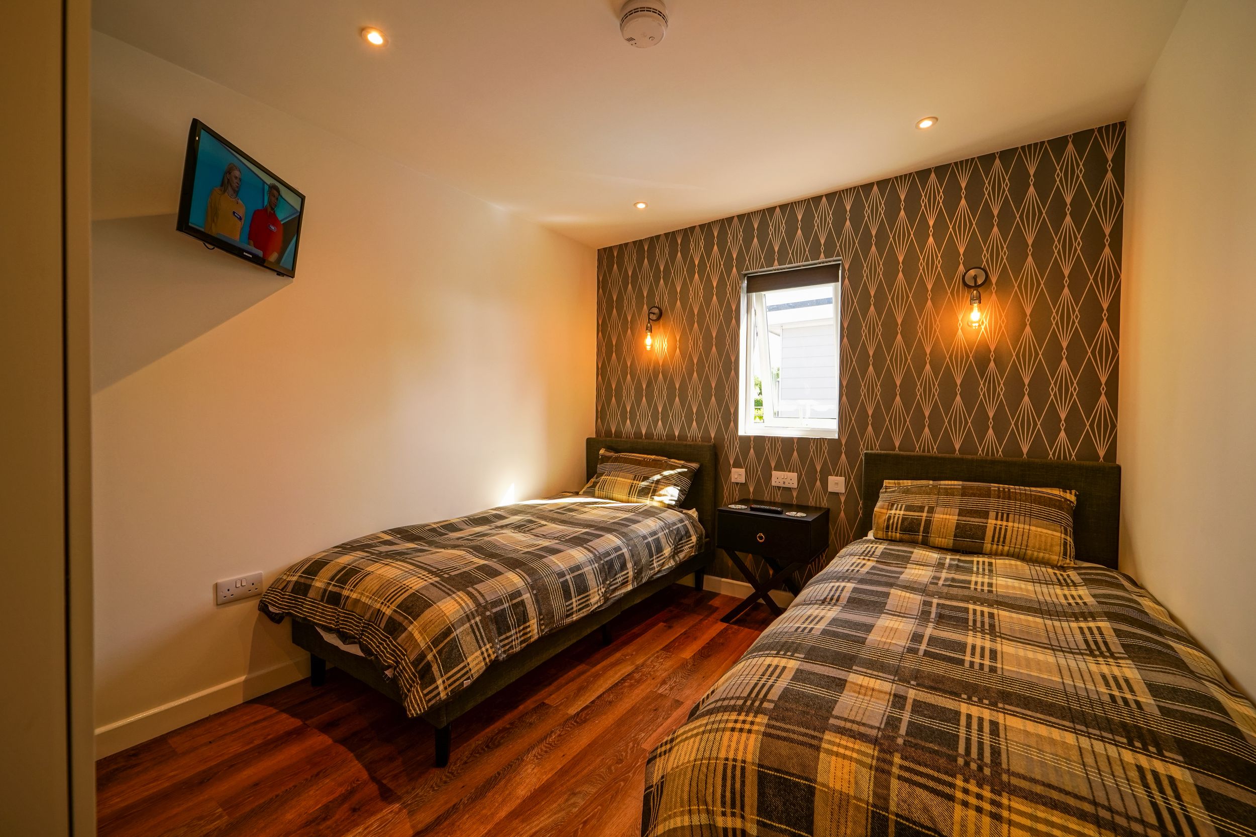Modern bedroom features two single beds with plaid bedding, wooden flooring, and a geometric wallpaper accent wall.