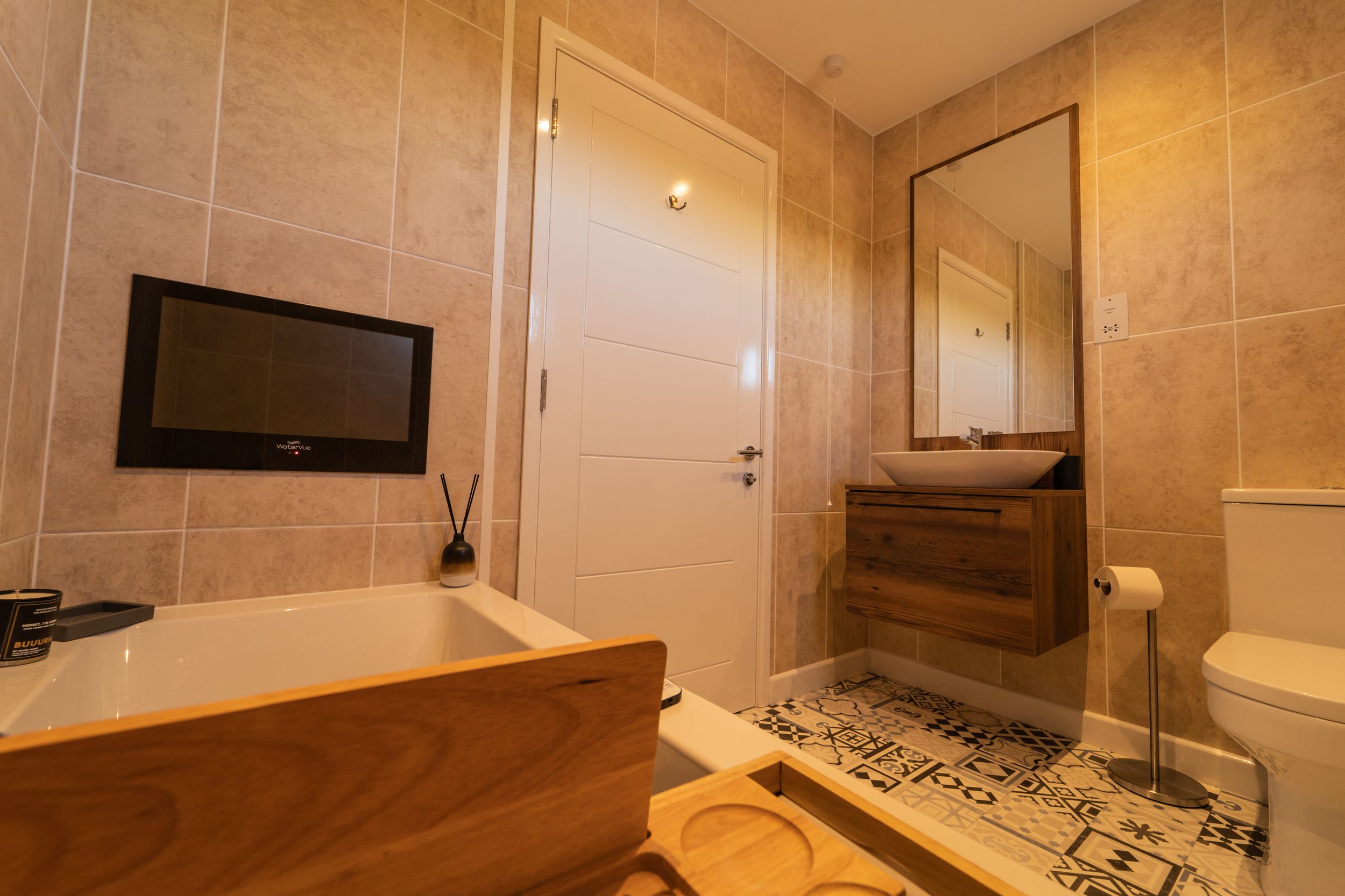 Modern bathroom interior with wooden bathtub, black-framed mirror, and geometric patterned floor tiles.