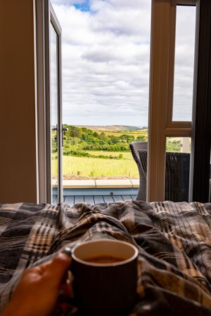 Cup of coffee on a plaid blanket by an open window overlooking rolling countryside hills on a cloudy day.