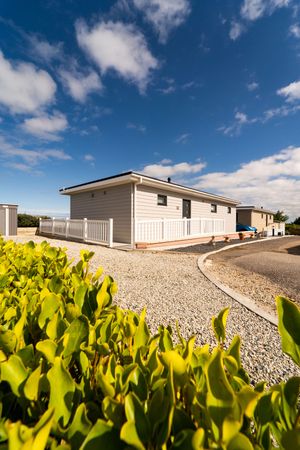 Gray and white mobile home with white railing deck viewed through bright green bushes under a blue cloudy sky.