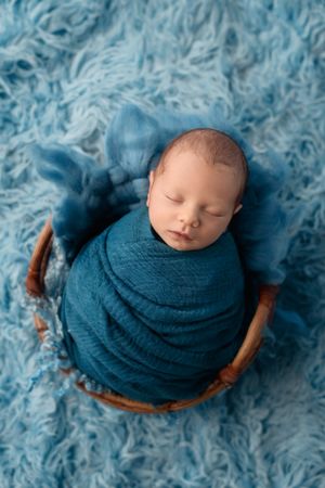 Newborn baby wrapped in a blue wrap in a rustic wooden basket on a fluffy blue rug