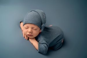 Newborn baby sleeping on blue soft backdrop with hands under chin with blue matching hat