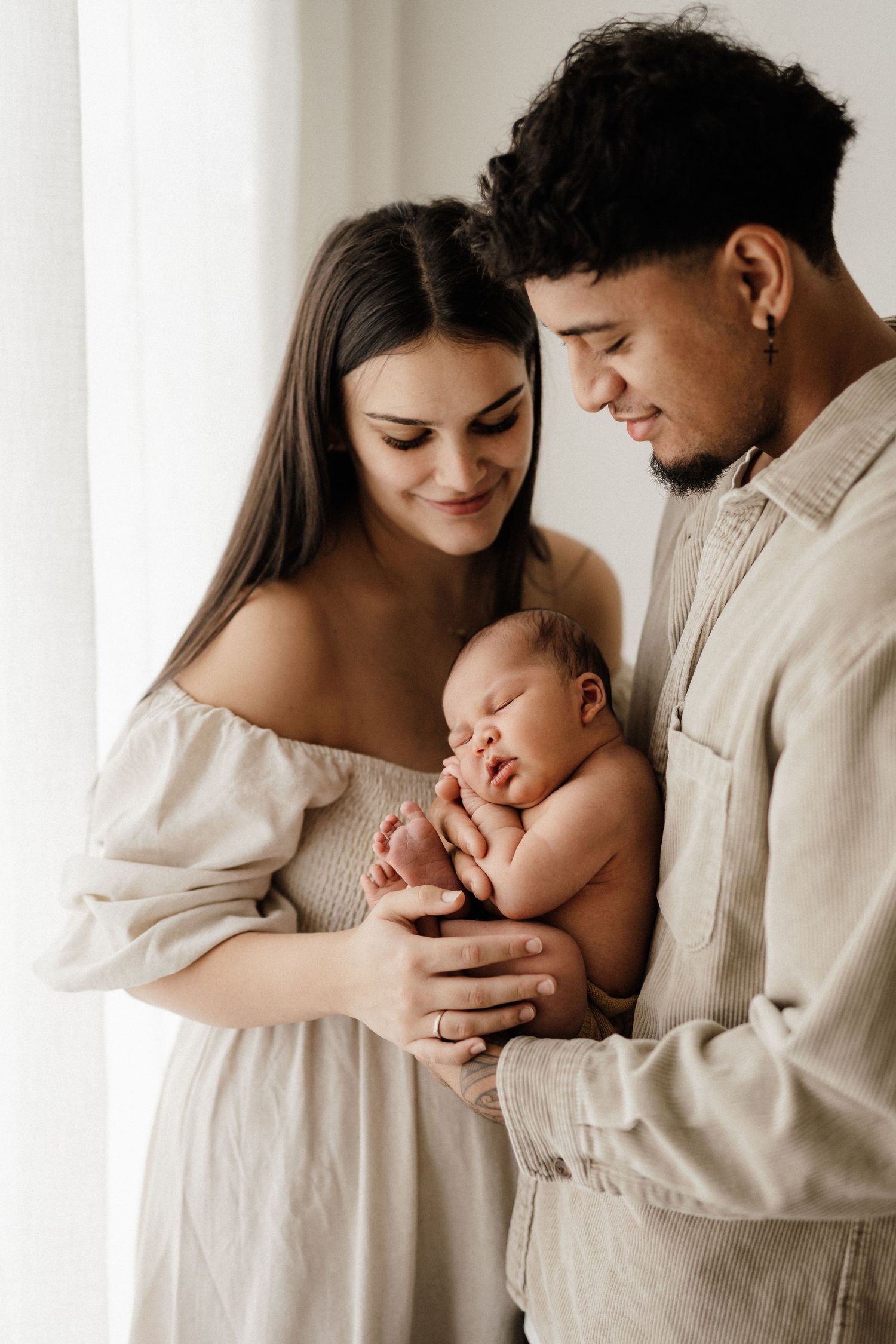 Parents sharing tender moment with sleeping newborn baby in white room.