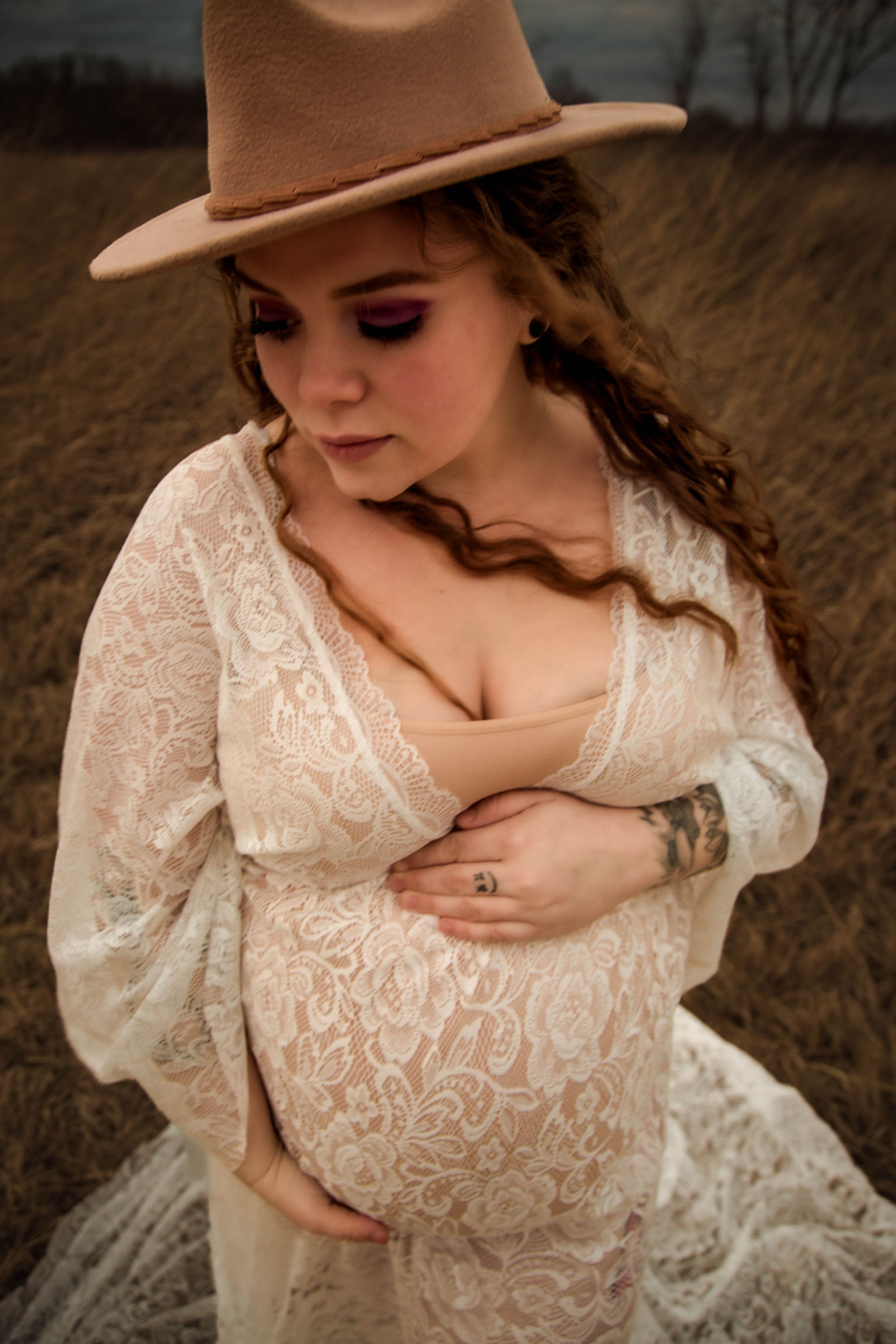 Pregnant woman in lace boho dress and tan wide-brim hat cradling her baby bump in a moody outdoor field.
