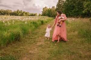 Mother in pink dress walking with toddler and baby through a wildflower meadow.