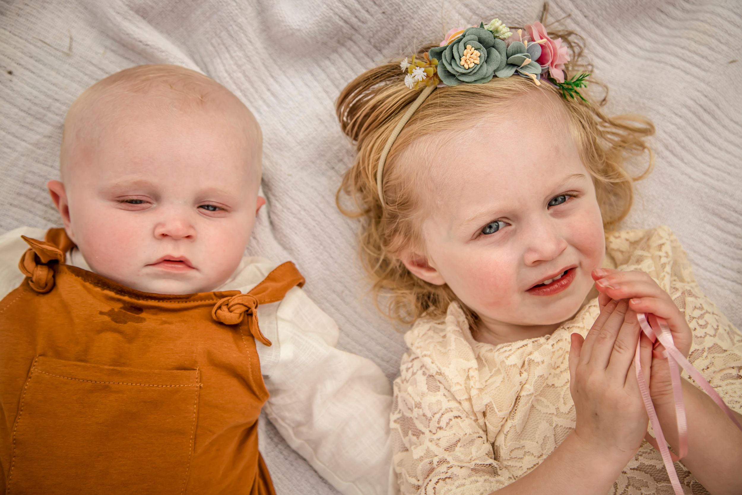 Two babies lying on a white blanket, one in orange romper and one in cream dress with floral headband.