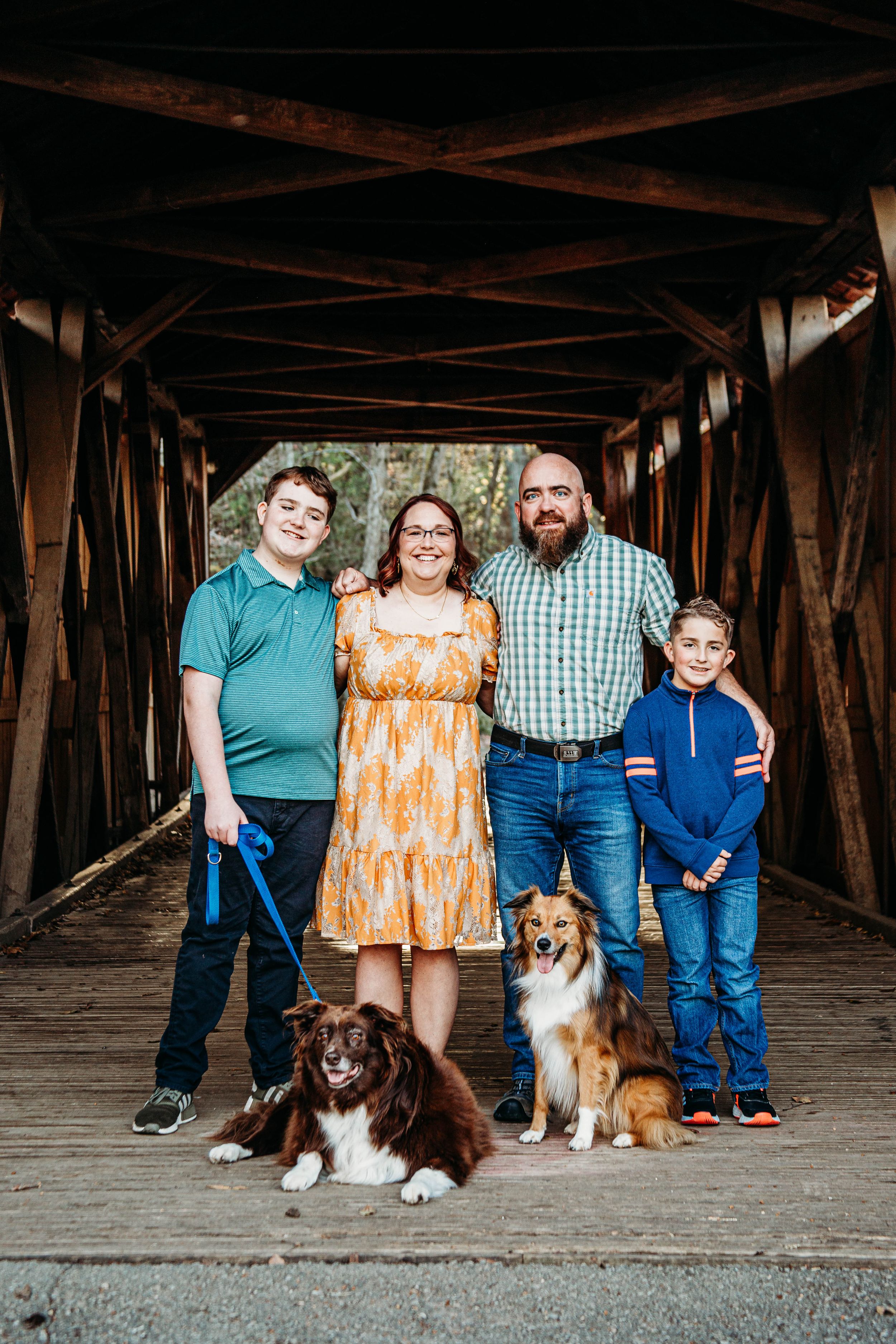 Family of four posing with two dogs on a wooden covered bridge.