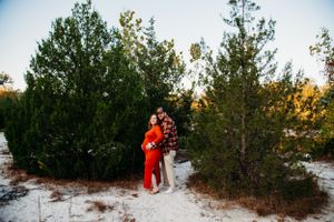 Pregnant couple embracing among evergreen trees on white sand at sunset.